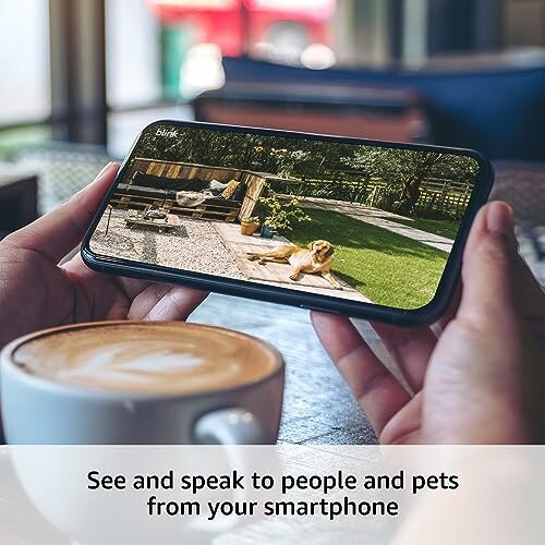 Person holding a smartphone showing a dog in a garden, with a coffee cup nearby.